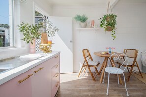 A kitchen with a white countertop, pink cabinets, and a window. There is a dining area with a round table, two chairs, and hanging plants. - Palm Springs Villa, Walk to Beaches & Free Parking (Bateau Bay)
