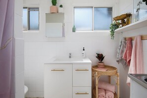 A bathroom with white tiles, a white sink, a small wicker stool, and a shelf with towels. - Palm Springs Villa, Walk to Beaches & Free Parking (Bateau Bay)