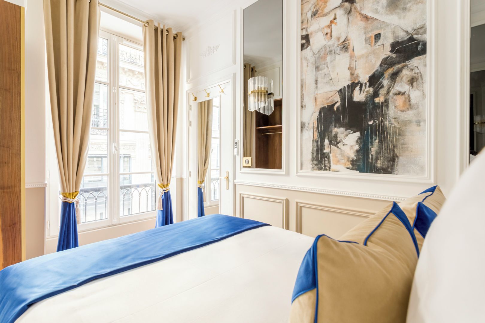Habitación de hotel con una cama grande, cortinas color beige, vistas a la ciudad y un cuadro abstracto moderno en la pared.
