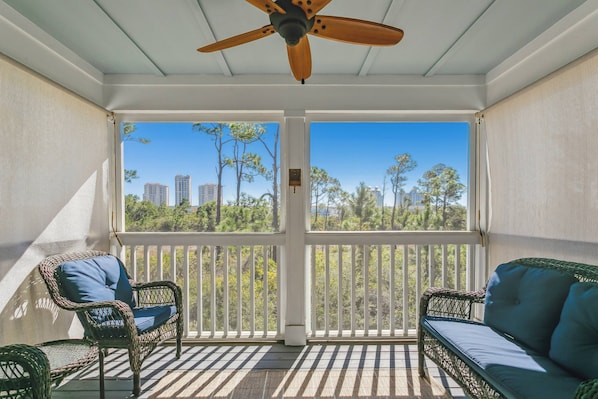 A covered balcony with wicker furniture, a ceiling fan, and a view of trees and buildings.