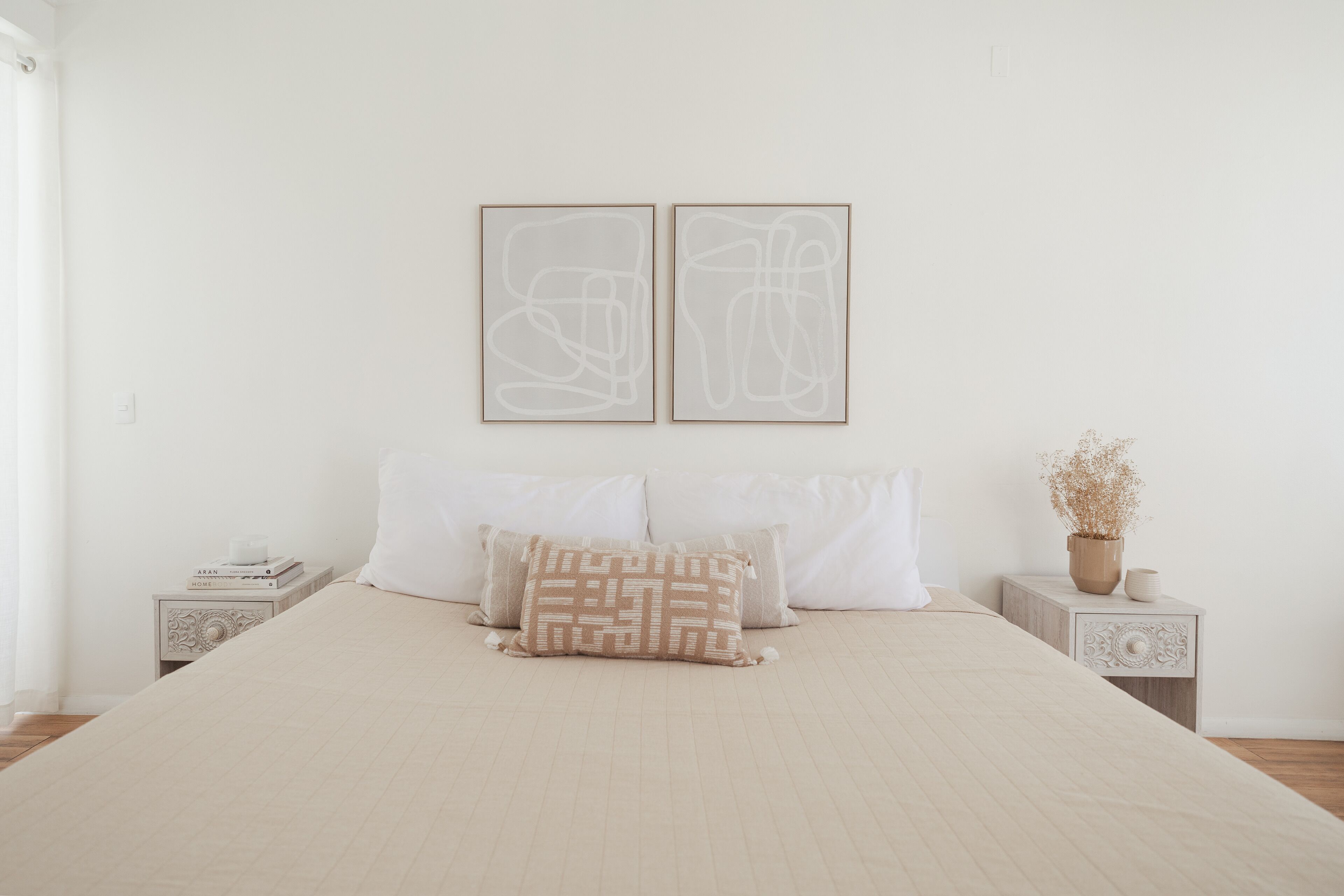 A neatly made bed with a beige bedspread and two pillows, a nightstand with a decorative item, and a wall with two abstract art pieces.