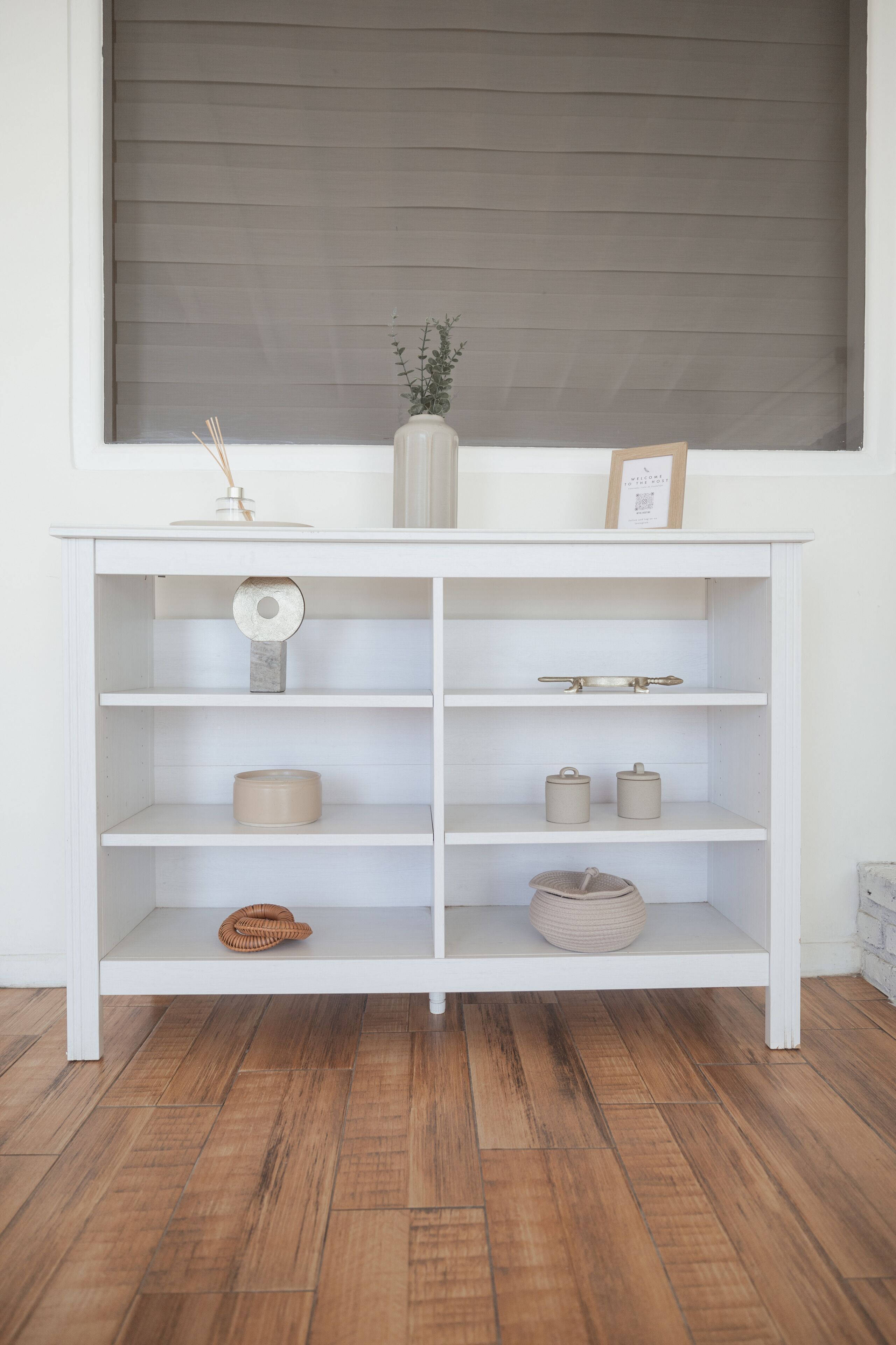 A white wooden shelf unit with various items on it, including a vase with a plant, a picture frame, and some containers.