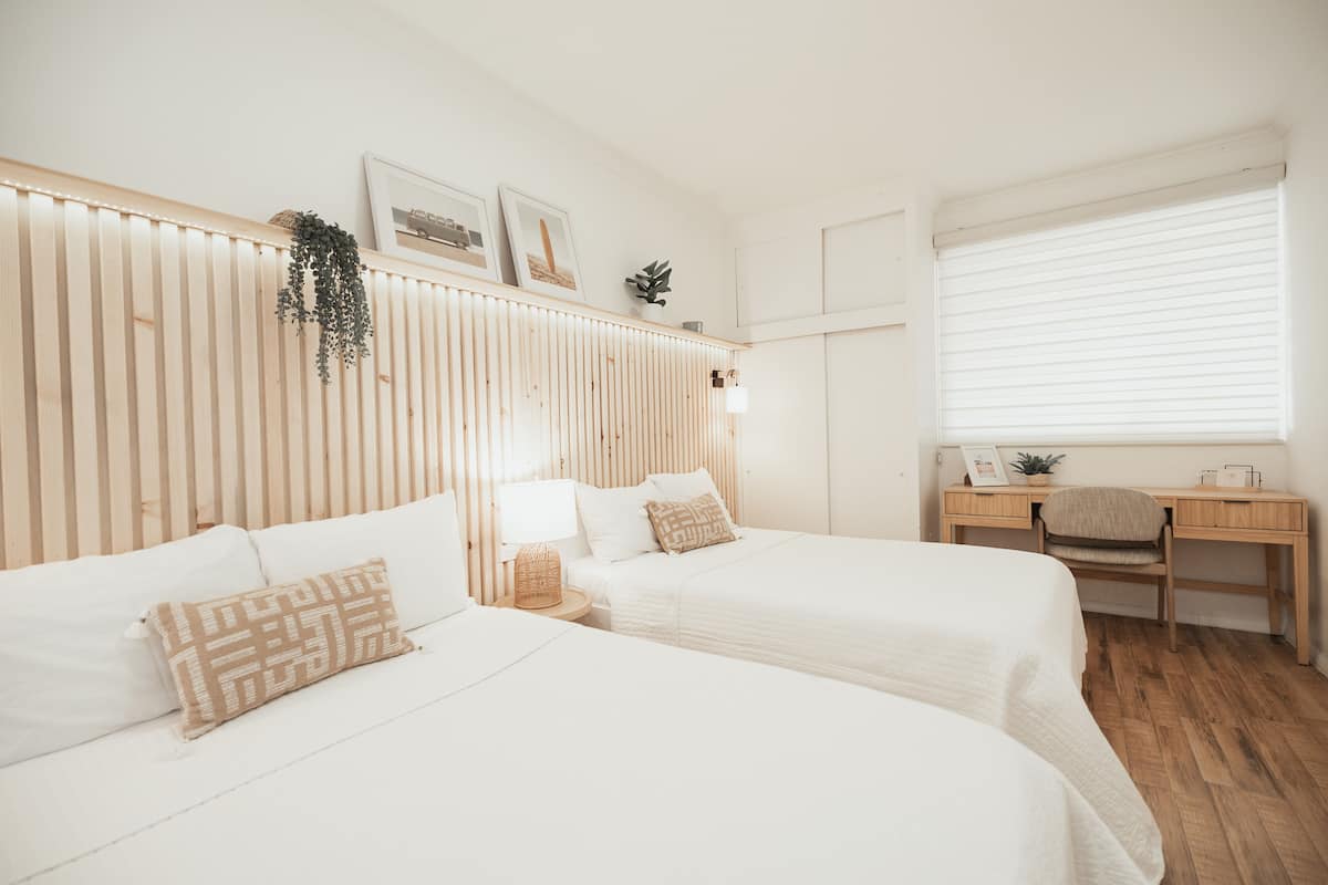 A hotel room with two beds, a wooden headboard, a desk with a chair, and a window with blinds.