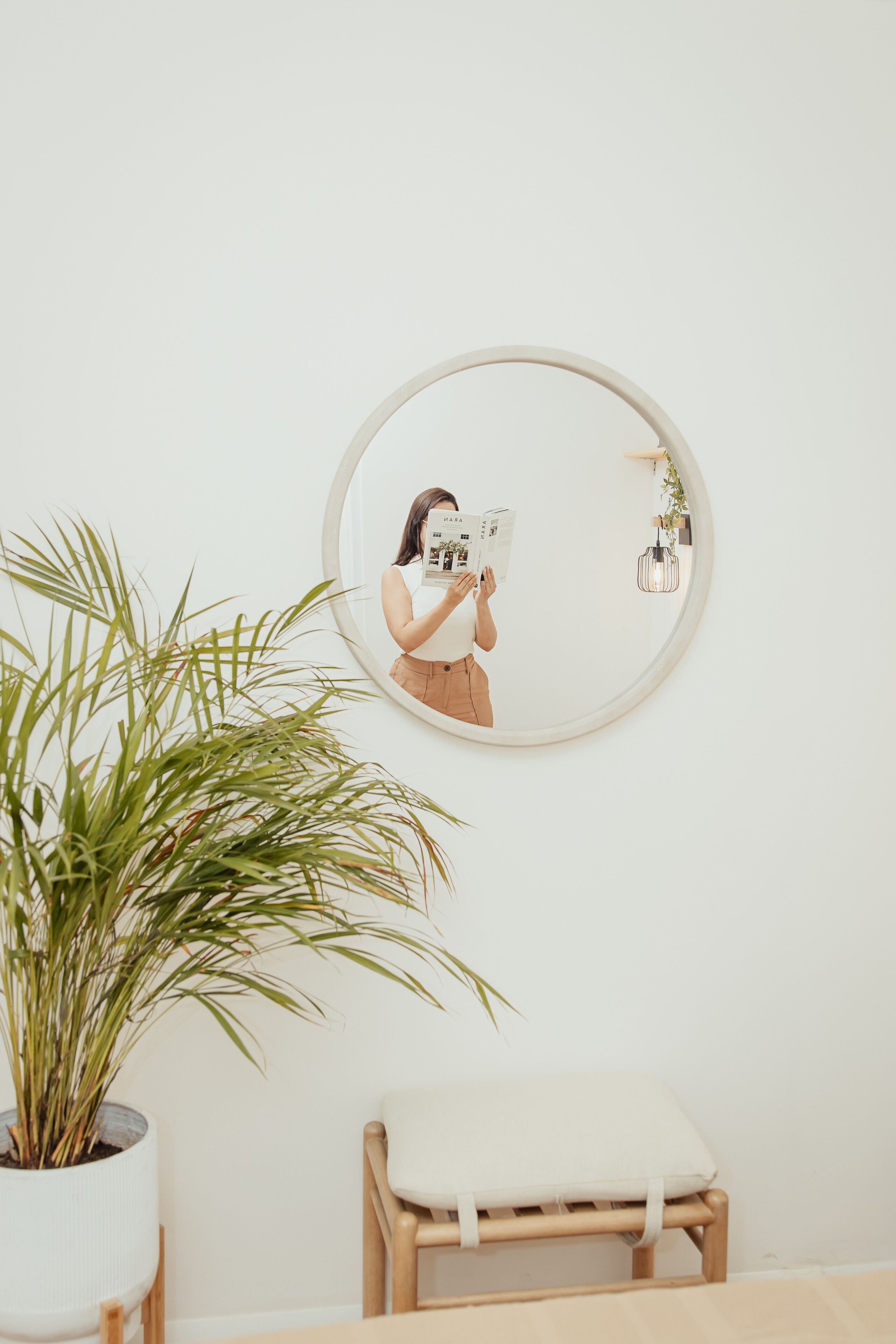 Une pièce avec un grand miroir rond au mur, une plante en pot et un petit tabouret en bois.