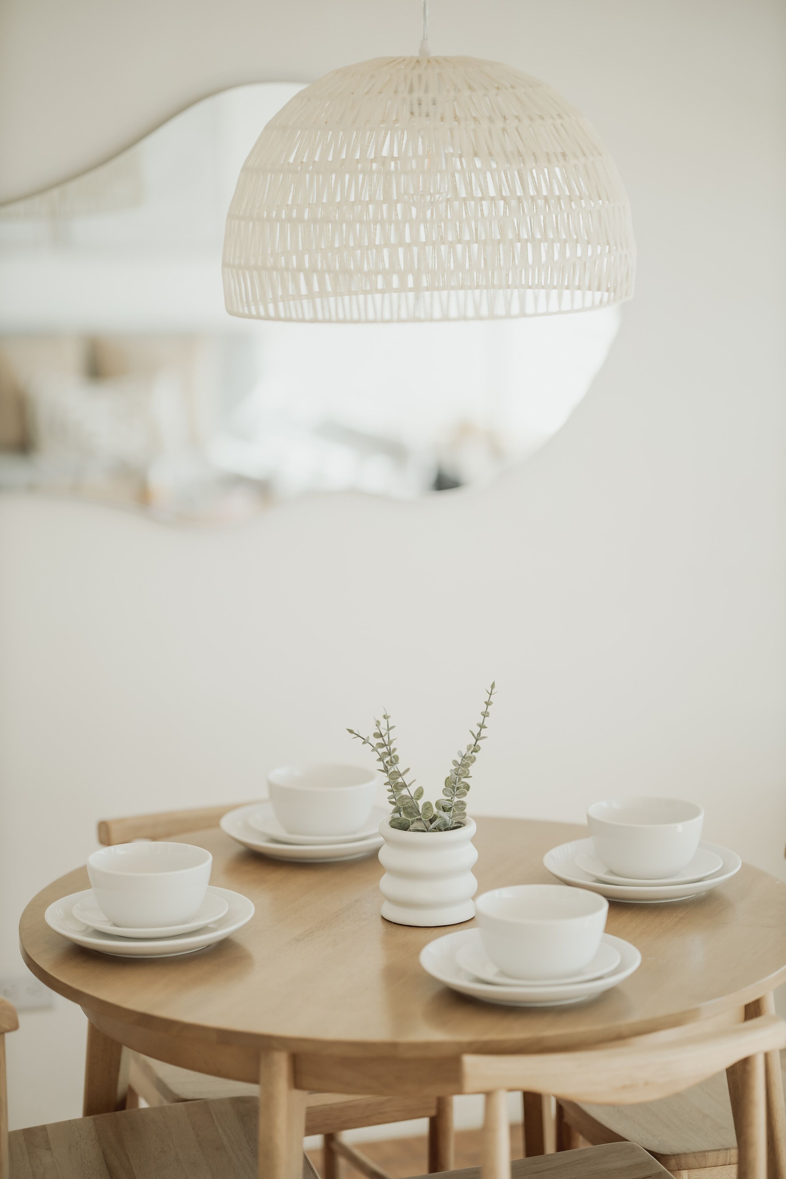 A round wooden table with white bowls and saucers, a small white vase with greenery, and a textured white pendant light.