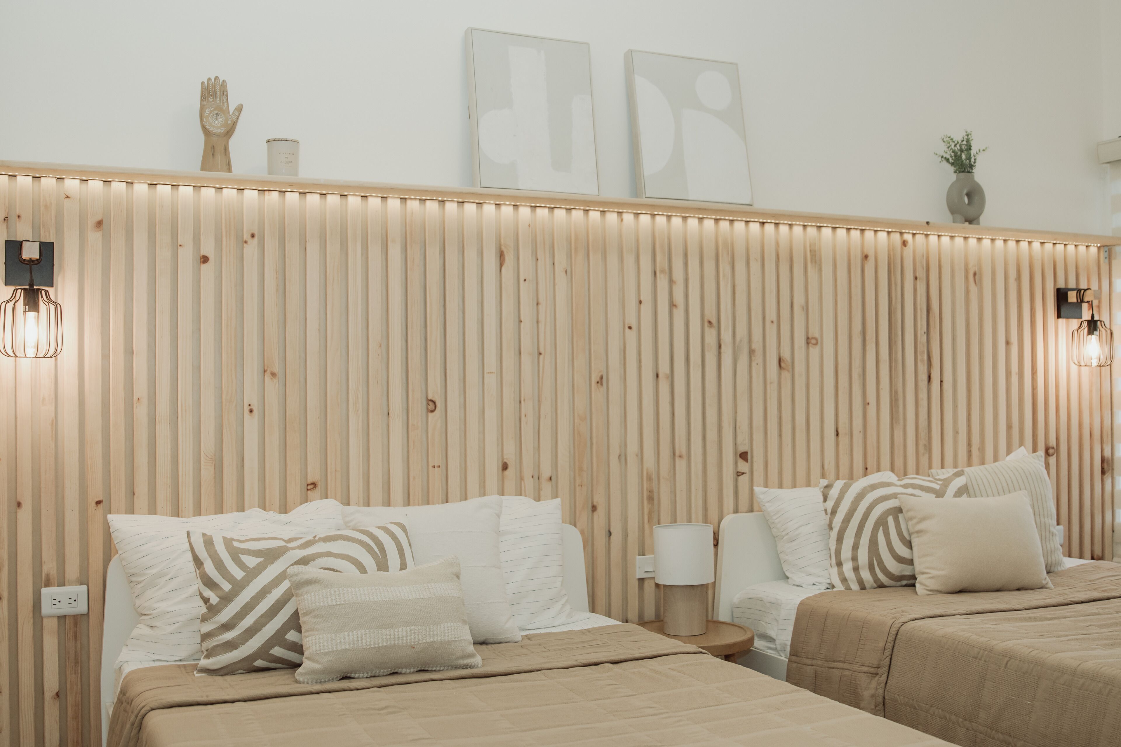 A hotel room with two beds, a wooden slat headboard, wall-mounted light fixtures, and a small side table.