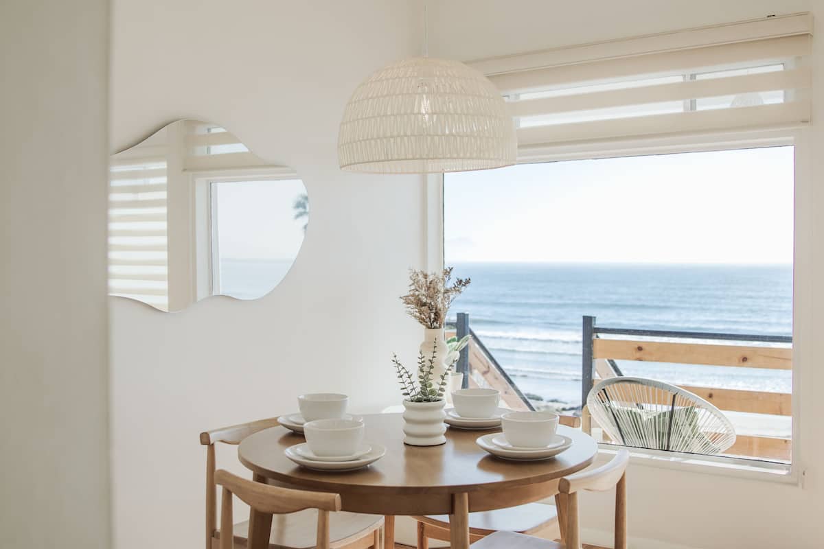 A dining area with a round table, white chairs, and a view of the ocean through large windows.