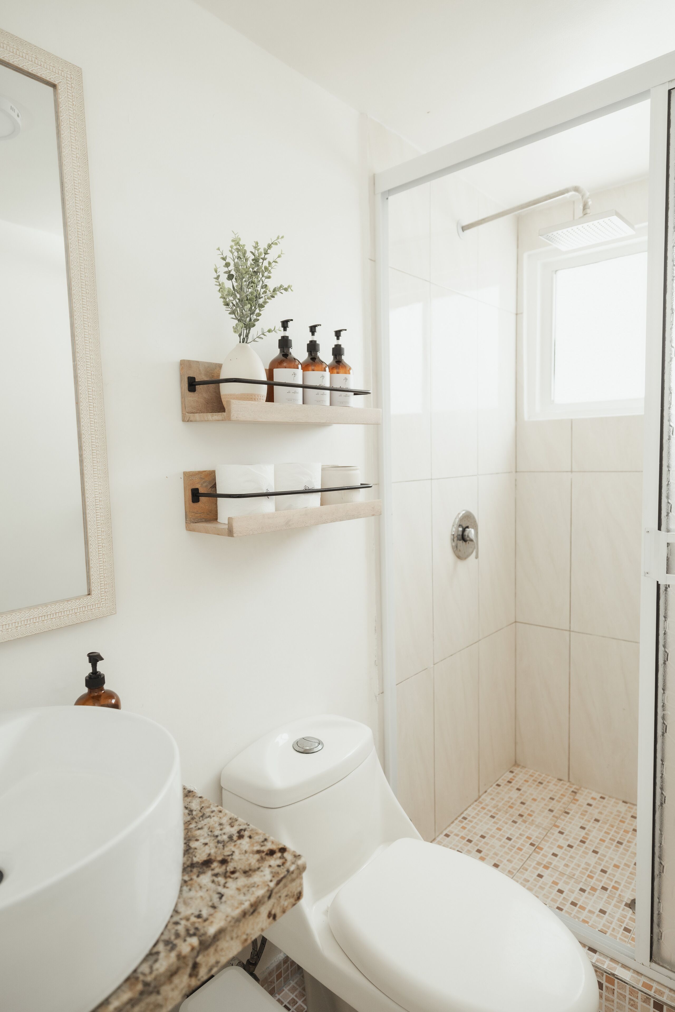 Une salle de bain avec un lavabo, des toilettes et une douche, le tout de couleur blanche. On y trouve des étagères en bois où sont rangés les articles de toilette, et un miroir encadré au-dessus du lavabo.