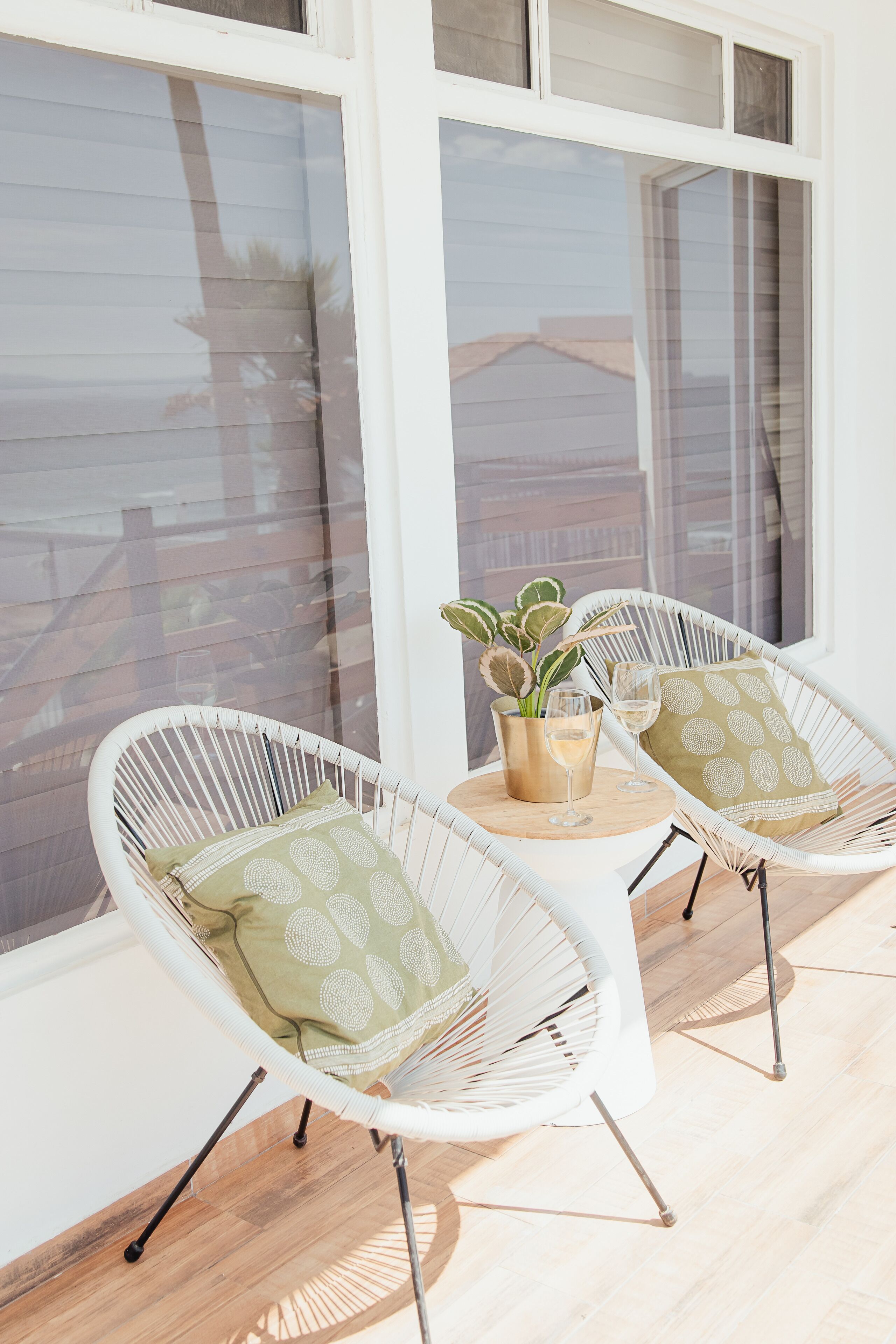 Two white wicker chairs with cushions, a small round table with a plant and two glasses, and a view of a building through large windows.