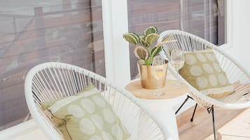Two white wicker chairs with cushions, a small round table with a plant and two glasses, and a view of a building through large windows.