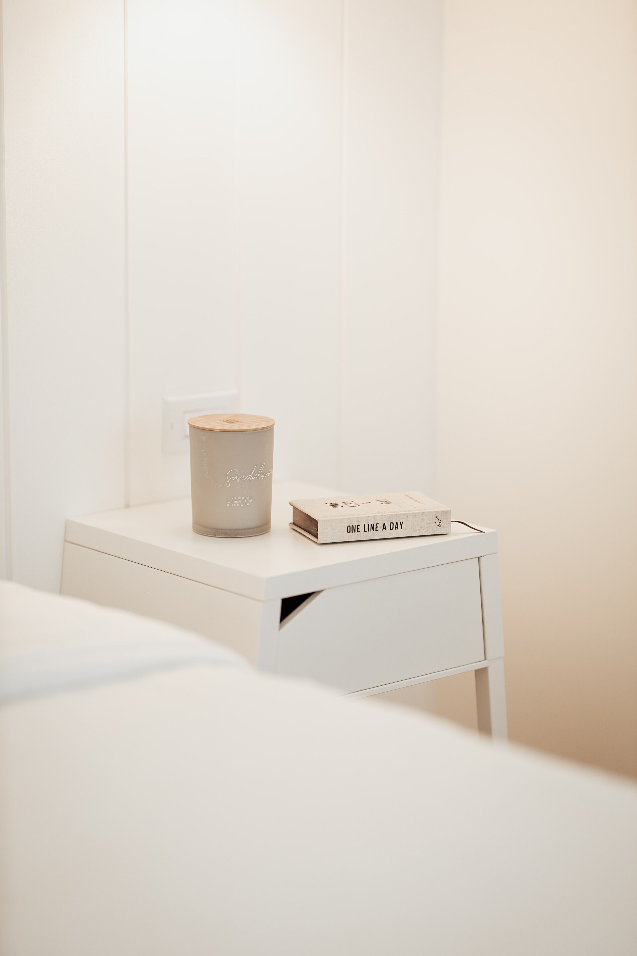 Una mesita de noche blanca con una vela y un libro titulado 'One Line a Day'.