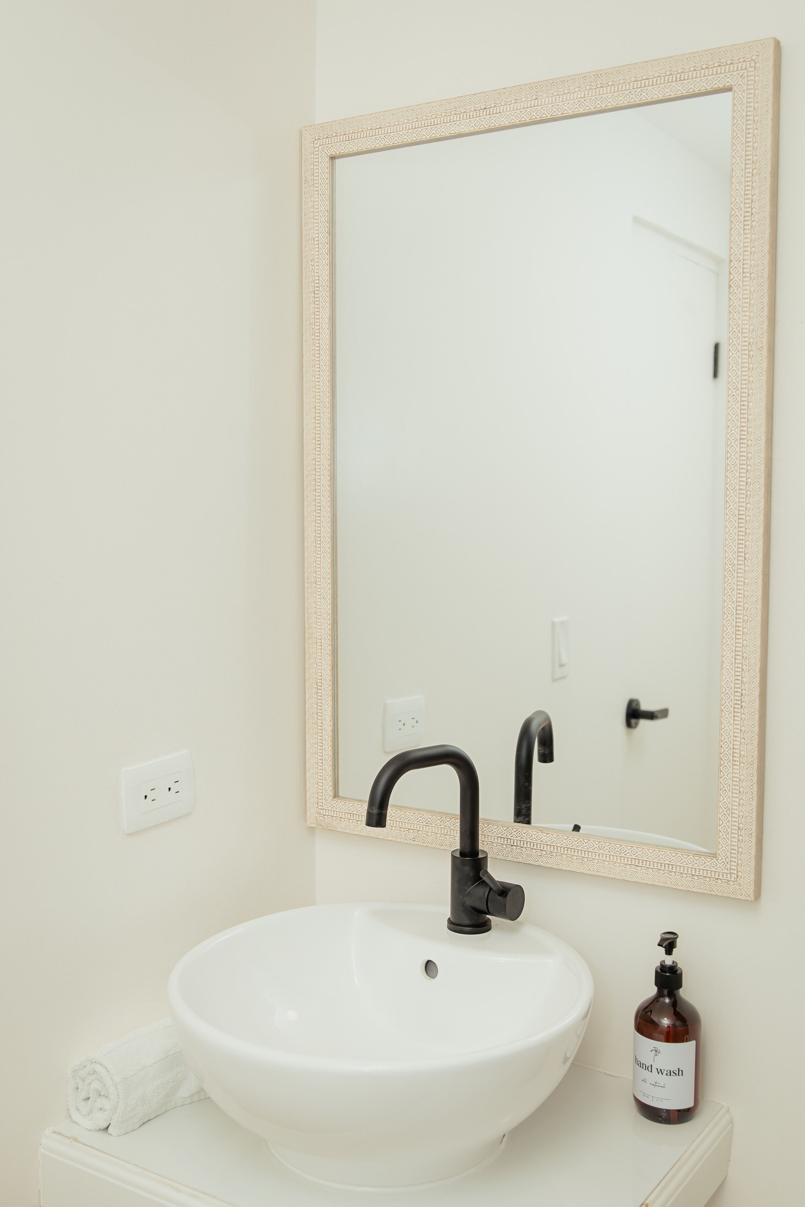 A bathroom with a white sink, a mirror with a decorative frame, a black faucet, a towel, and a bottle of hand wash soap.