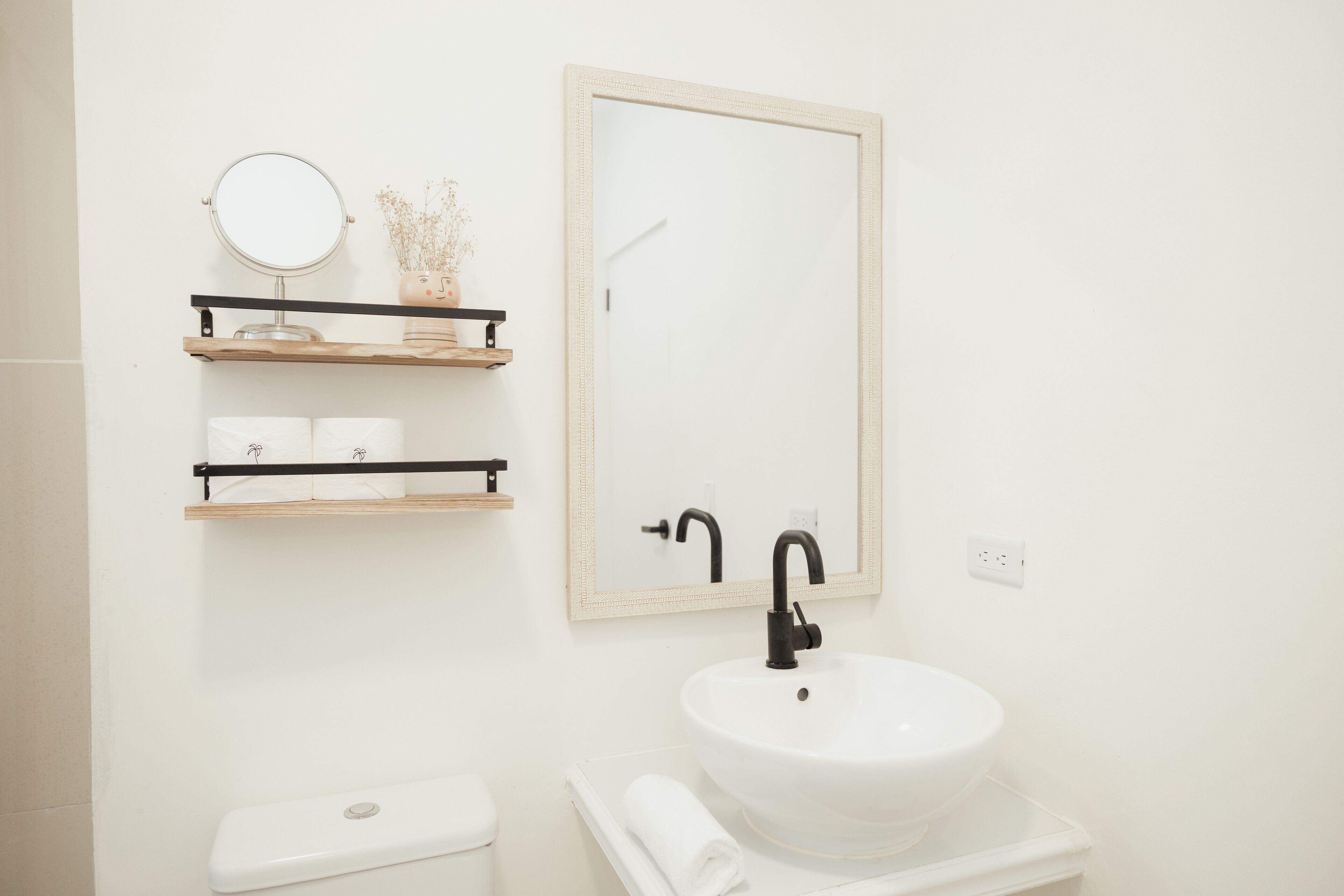 Een badkamer met een wit wastafel, een zwarte kraan en een spiegel erboven. Er zijn twee planken met toiletartikelen en een toilet zichtbaar.