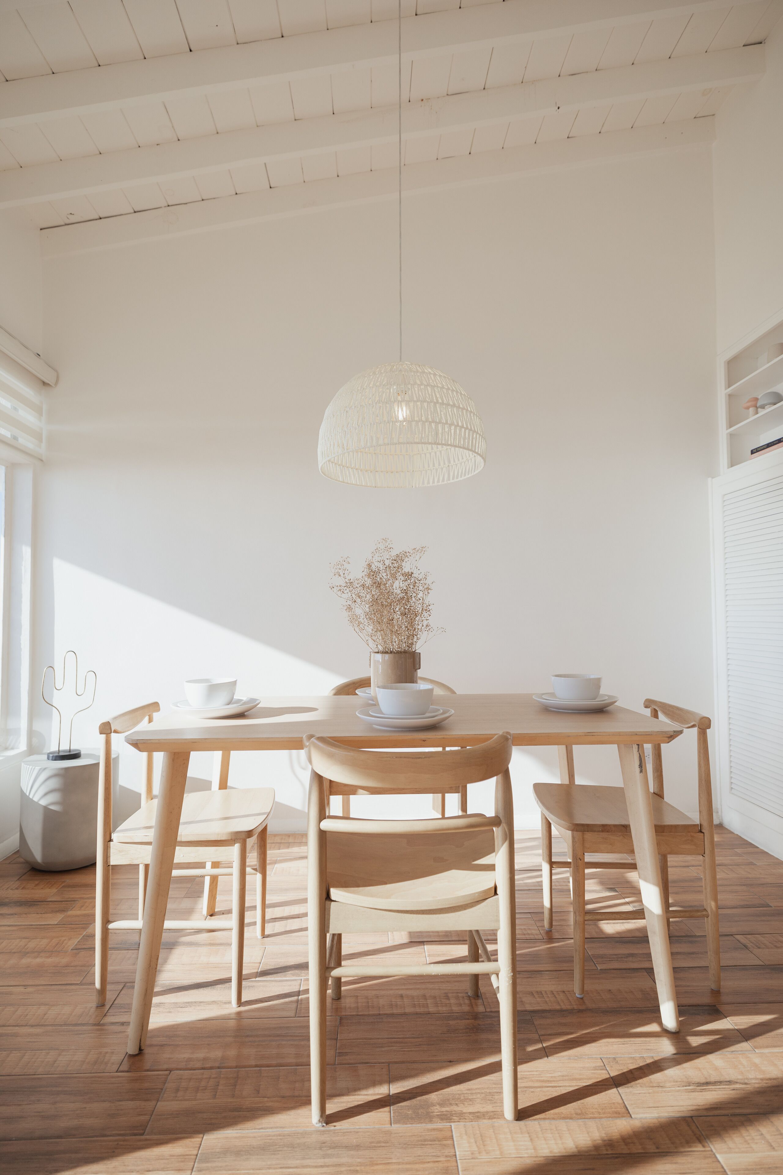 Un coin repas avec une table et des chaises en bois, un luminaire suspendu et un vase contenant des fleurs séchées.