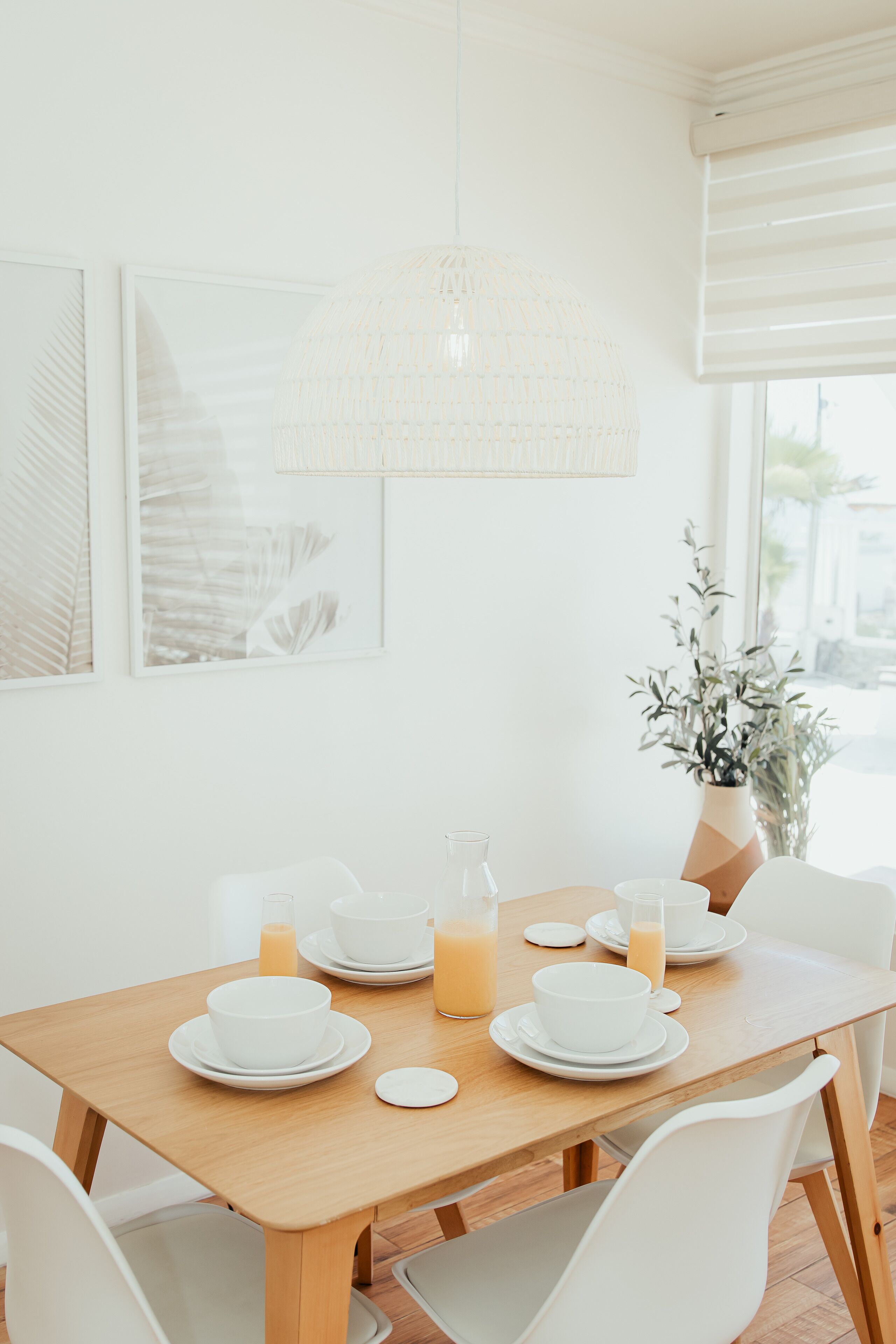 A dining area with a wooden table set for breakfast, white chairs, a hanging light fixture, and a window with blinds.