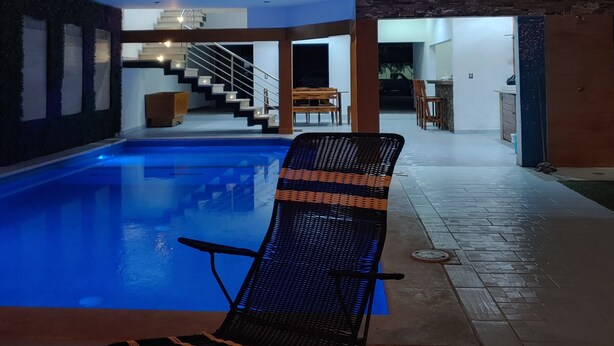 A modern indoor swimming pool with a lounge chair, a staircase leading to an upper level, and a stone wall.