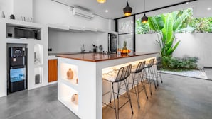 A modern kitchen with a central island, bar stools, and a large window overlooking a garden. - Sunny Home Villa (Canggu)