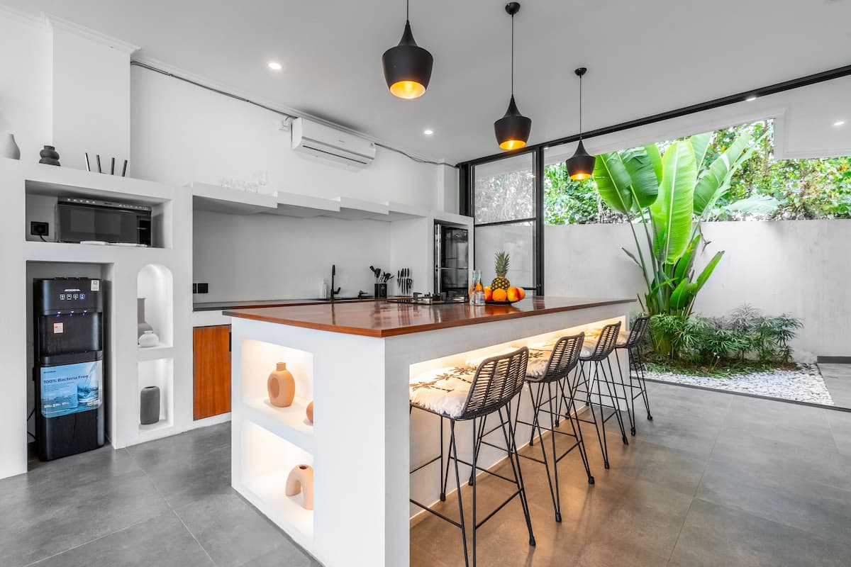 A modern kitchen with a central island, bar stools, and a large window overlooking a garden.