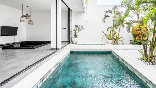 A modern indoor pool area with a clear pool, a seating area with black cushions, and potted plants around the pool. - Sunny Home Villa (Canggu)