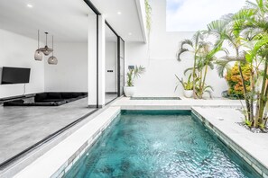 A modern indoor pool area with a clear pool, a seating area with black cushions, and potted plants around the pool. - Sunny Home Villa (Canggu)