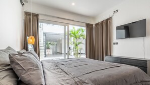 A modern bedroom with a large bed, a flat-screen TV, and a sliding glass door leading to a balcony with a plant. - Sunny Home Villa (Canggu)