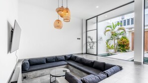 A modern living room with a sectional sofa, a small coffee table, and a TV mounted on the wall. There are three hanging wicker lamps and a large window providing a view of the outdoor area with plants. - Sunny Home Villa (Canggu)