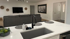 A modern kitchen with a large sink, a dining area with a grey sofa, and a flat-screen TV on the wall. - Quiet 3BR Near Hospital & Georgia Southern | 2 King Beds + Workspace (Statesboro)