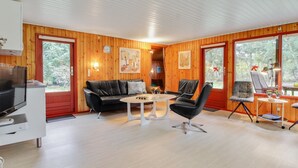 Een knus blokhutinterieur met houten afwerking, een zwarte leren bank, een ronde tafel en een tv op een standaard.