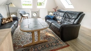 A modern living room with a black leather sofa, a round wooden table, and a patterned rug. There are two windows providing natural light and a view of greenery outside. - "Arnora" - 48km from the sea by Interhome (Højer)