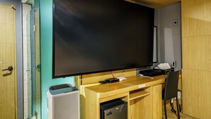 Habitación con televisor de pantalla plana grande montado sobre un escritorio de madera, cabina de ducha de vidrio y un refrigerador compacto.