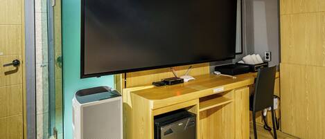 Una stanza con una grande TV a schermo piatto montata su una scrivania in legno, un box doccia in vetro e un frigorifero compatto.