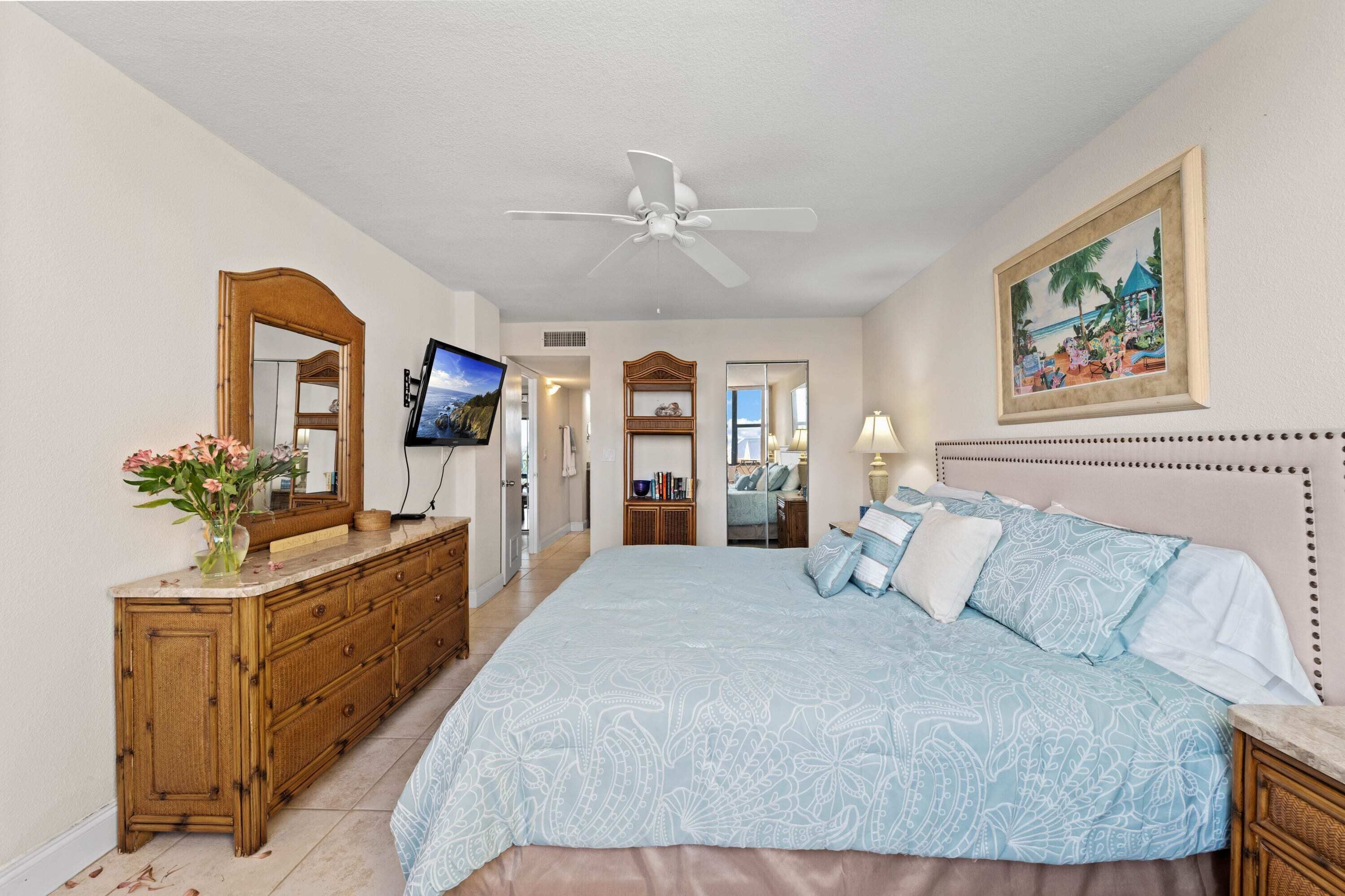 A bedroom with a large bed, a ceiling fan, a wooden dresser, a TV, and a framed picture on the wall.