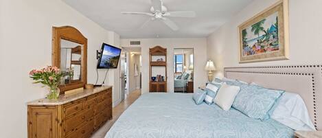 A bedroom with a large bed, a ceiling fan, a wooden dresser, a TV, and a framed picture on the wall.