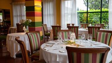 Un restaurante con mesas redondas dispuestas para comer, sillas a rayas y vistas a la vegetación a través de amplias ventanas.