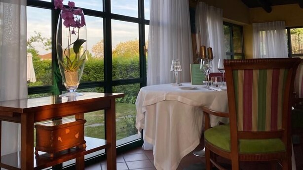 A dining area with a table set for two, large windows with curtains, and a view of greenery outside. - i castagni (Vigevano)
