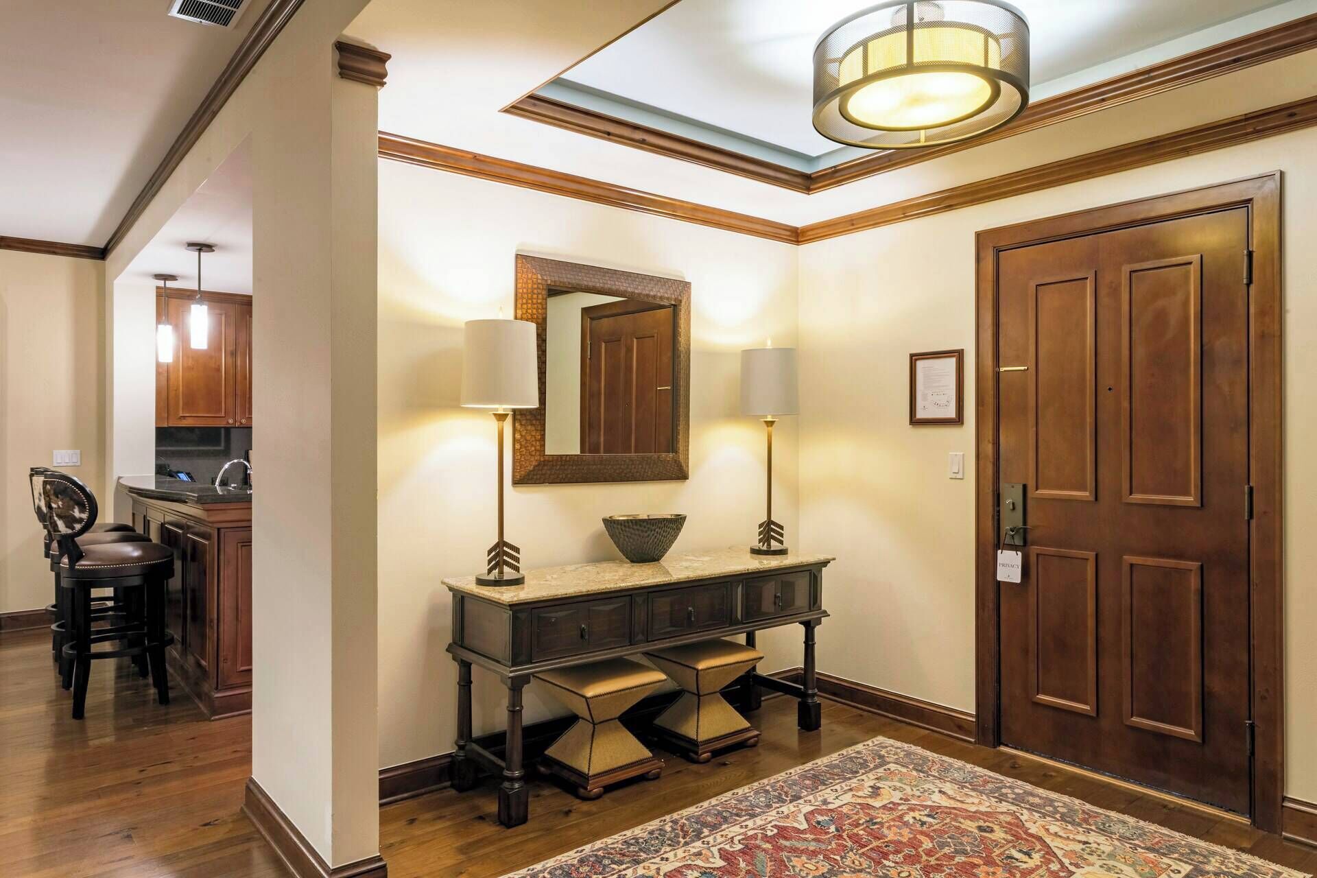 A well-lit hallway with a wooden door, a console table with lamps, a mirror, and a patterned rug.