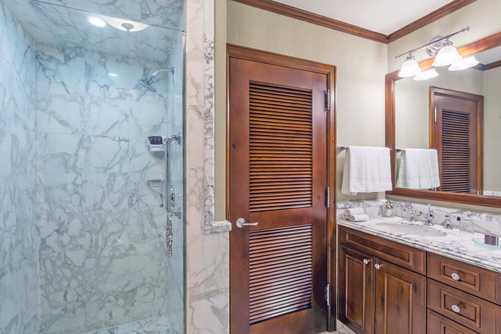 A bathroom with a marble shower, wooden door, vanity with a mirror, and a sink.