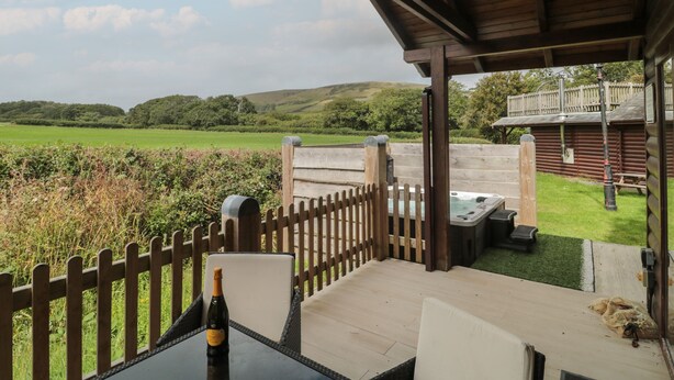 A covered patio with a table and chairs, a bottle of champagne, and a hot tub. - Oaks (Swanage)