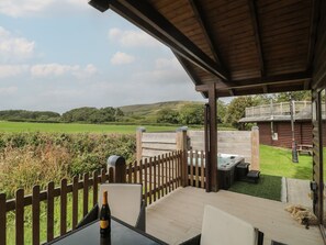 A covered patio with a table and chairs, a bottle of champagne, and a hot tub. - Oaks (Swanage)