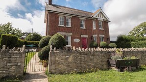 A two-story brick house with a red door, stone wall, and a garden with various plants and flowers. - The Farmhouse (Swanage)