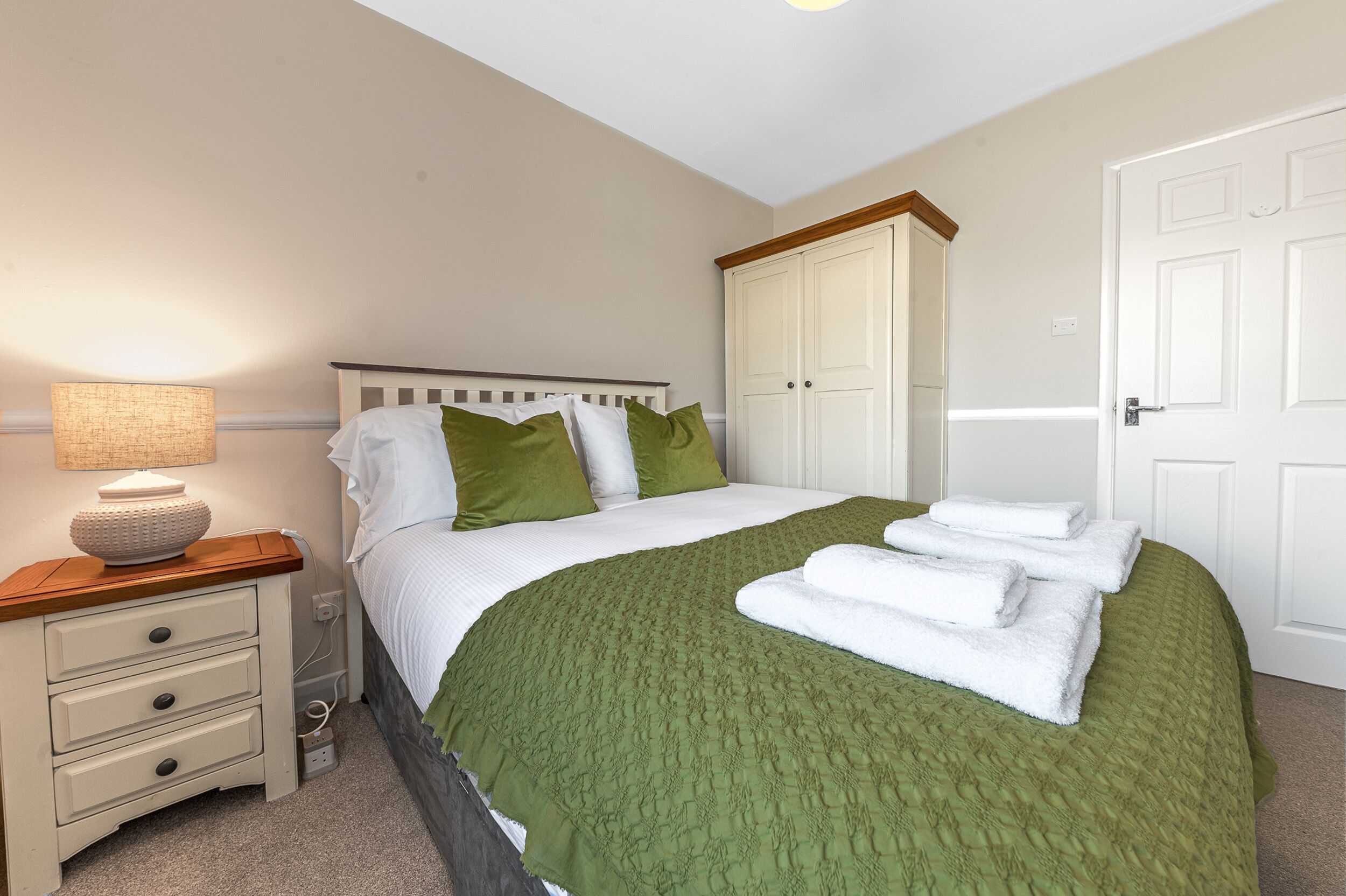 A neatly made bed with green and white bedding, a bedside lamp, and a wooden nightstand with drawers.