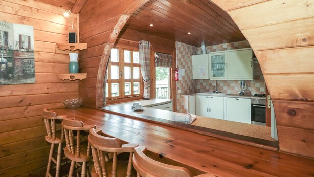 Un interior de madera con una zona de bar, sillas de madera, una cocina con armarios blancos y un ventanal grande.