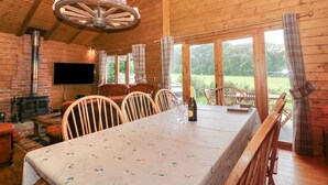 Interior de una cabaña de madera con una mesa de comedor, sillas, chimenea y ventanas amplias.