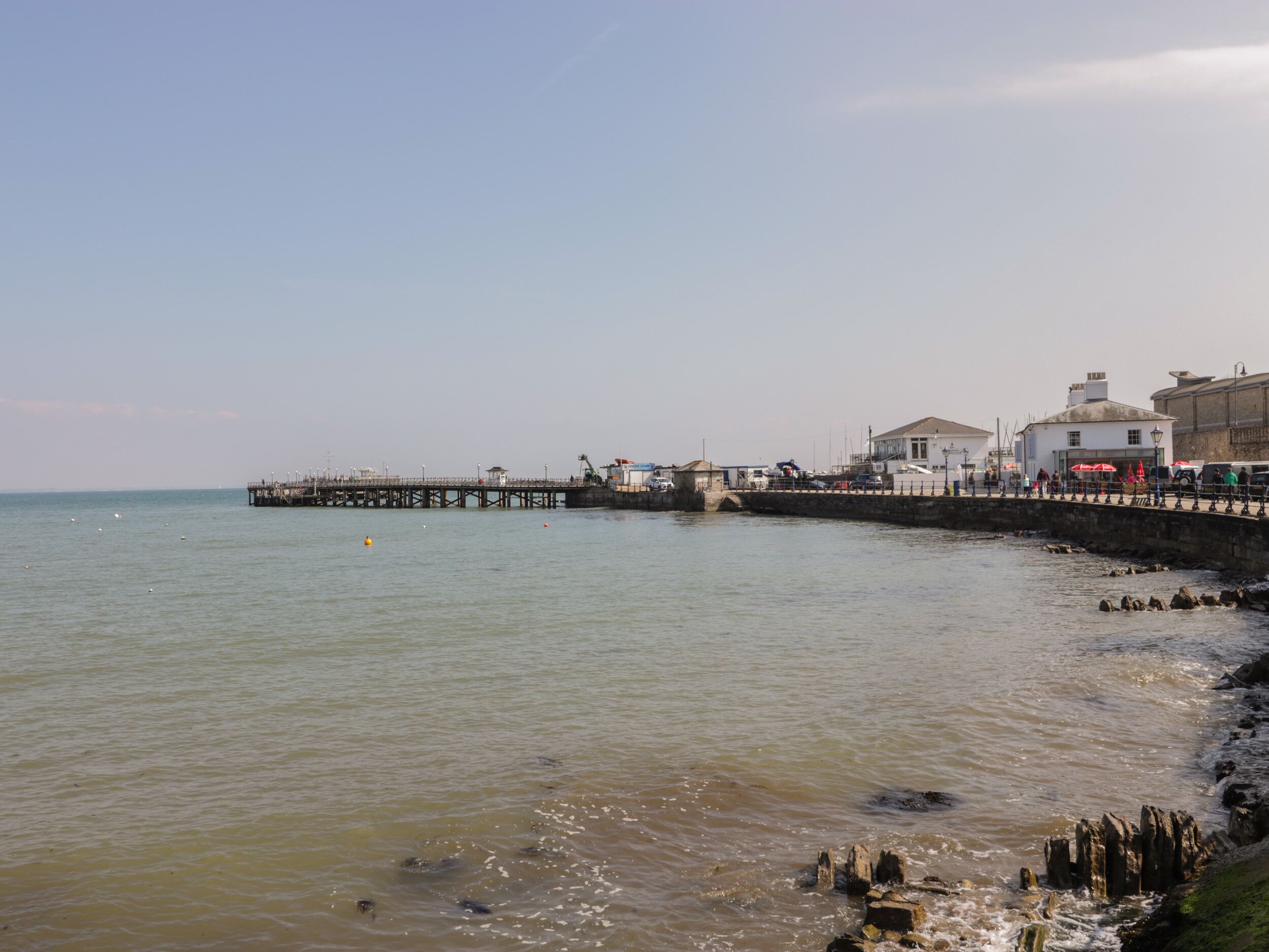 A coastal scene with a pier extending into the water, a stone wall along the shore, and buildings with visible windows and a flag.