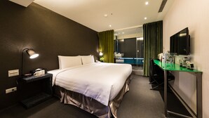 Quarto de hotel com uma cama grande, uma mesa com computador, televisão e banheiro visível através de uma porta de vidro.