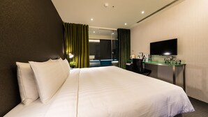 Quarto de hotel com uma cama grande, uma escrivaninha com televisão de tela plana e casa de banho visível através de uma porta de vidro.