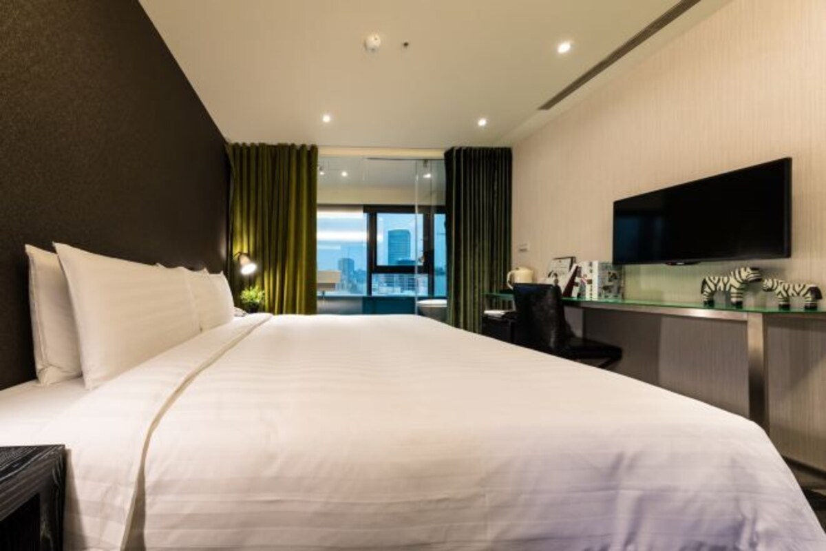 A hotel room with a large bed, a desk with a flat-screen TV, and a view of the city through the window.
