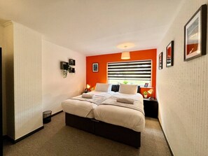 A bedroom with a bed, a nightstand, a wall-mounted lamp, and a window with blinds.