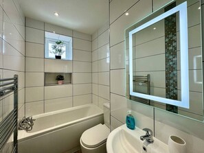 Un baño moderno con bañera, inodoro y lavamanos, con azulejos blancos y un espejo con marco de vidrio.