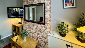 A living room with a brick fireplace, a mounted TV, a framed picture, and a small table with a plant. - Waterloo House – Contractors, Families, Holidays (Nottingham)